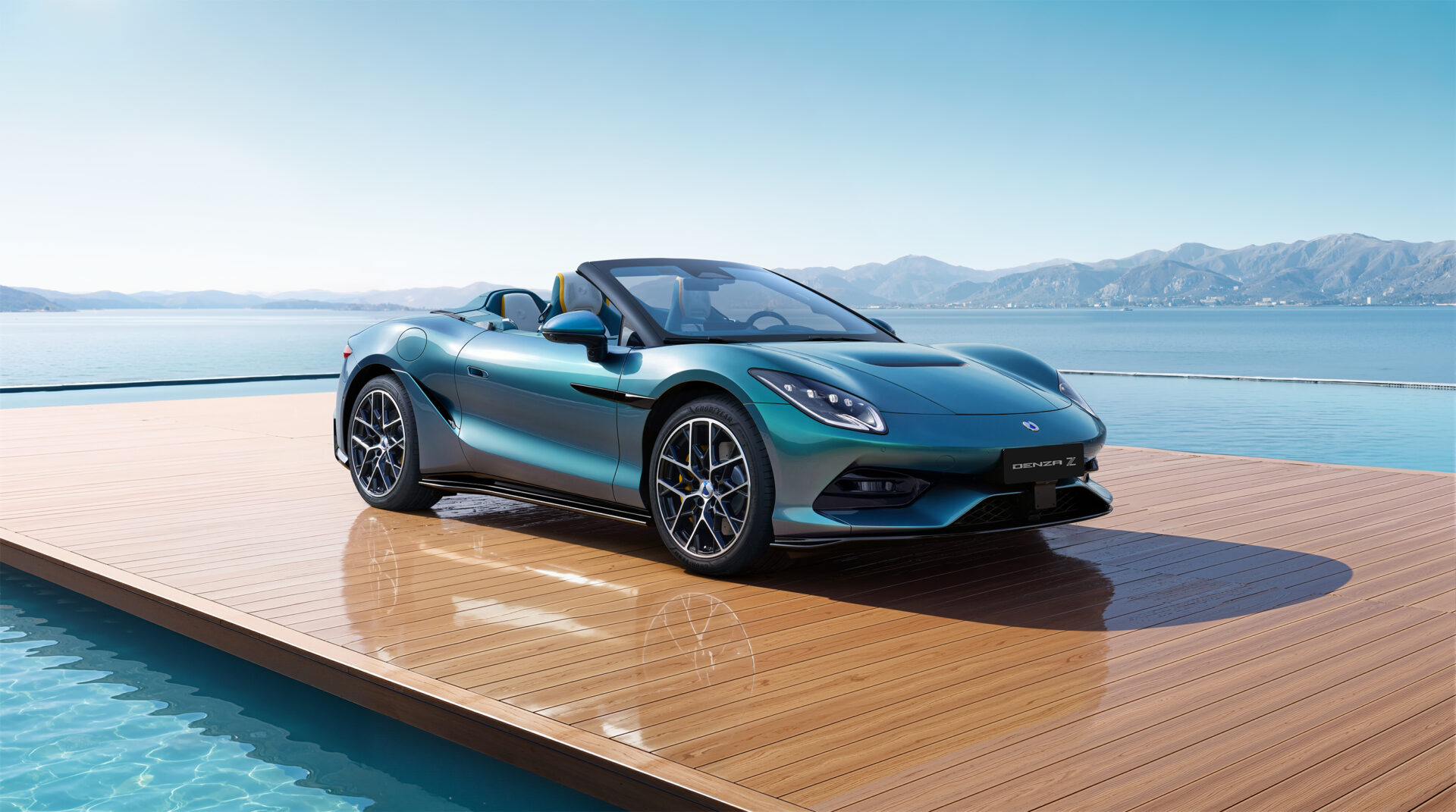 A sleek convertible car parked on a wooden deck by a body of water, with mountains in the background and clear blue skies.