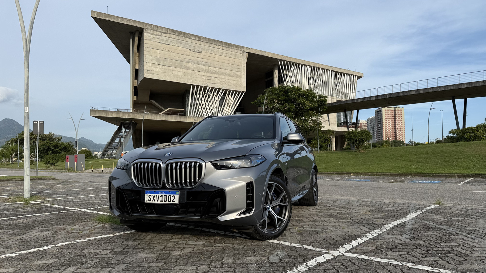 A silver BMW X5 parked in front of a modern architectural building, with green grass and mountains in the background.