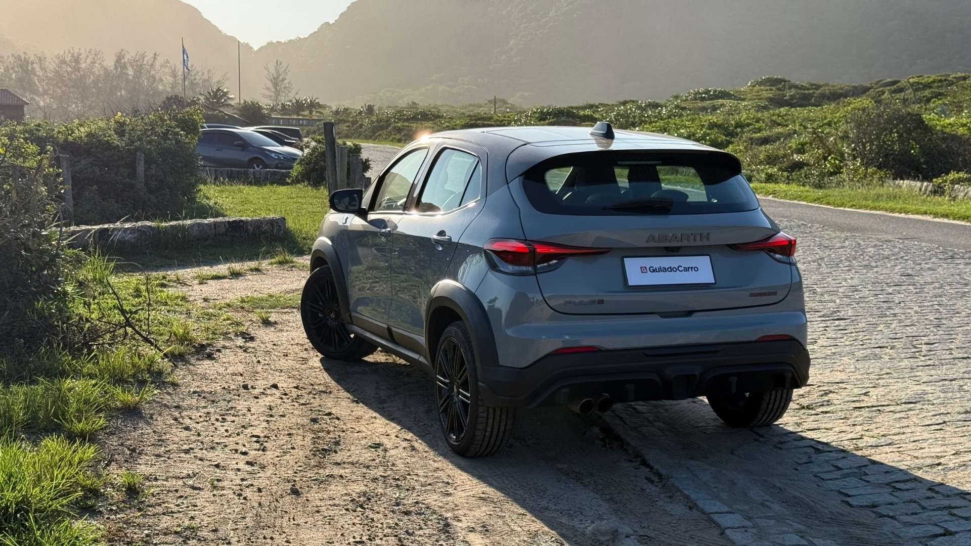 A silver Abarth SUV parked on the side of a cobblestone road, surrounded by greenery and mountains in the background, with sunlight shining on the vehicle.