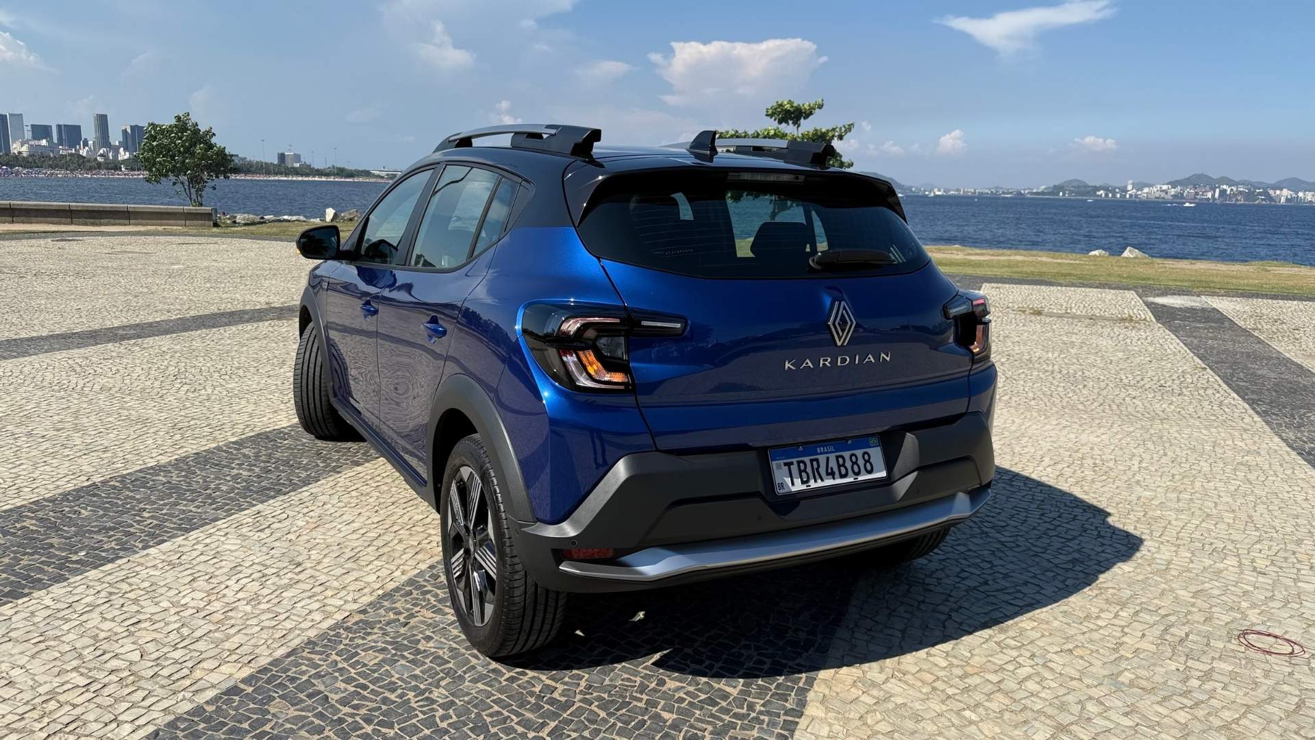 A blue Renault Kardian parked by the waterfront, showcasing its rear view with a city skyline in the background.