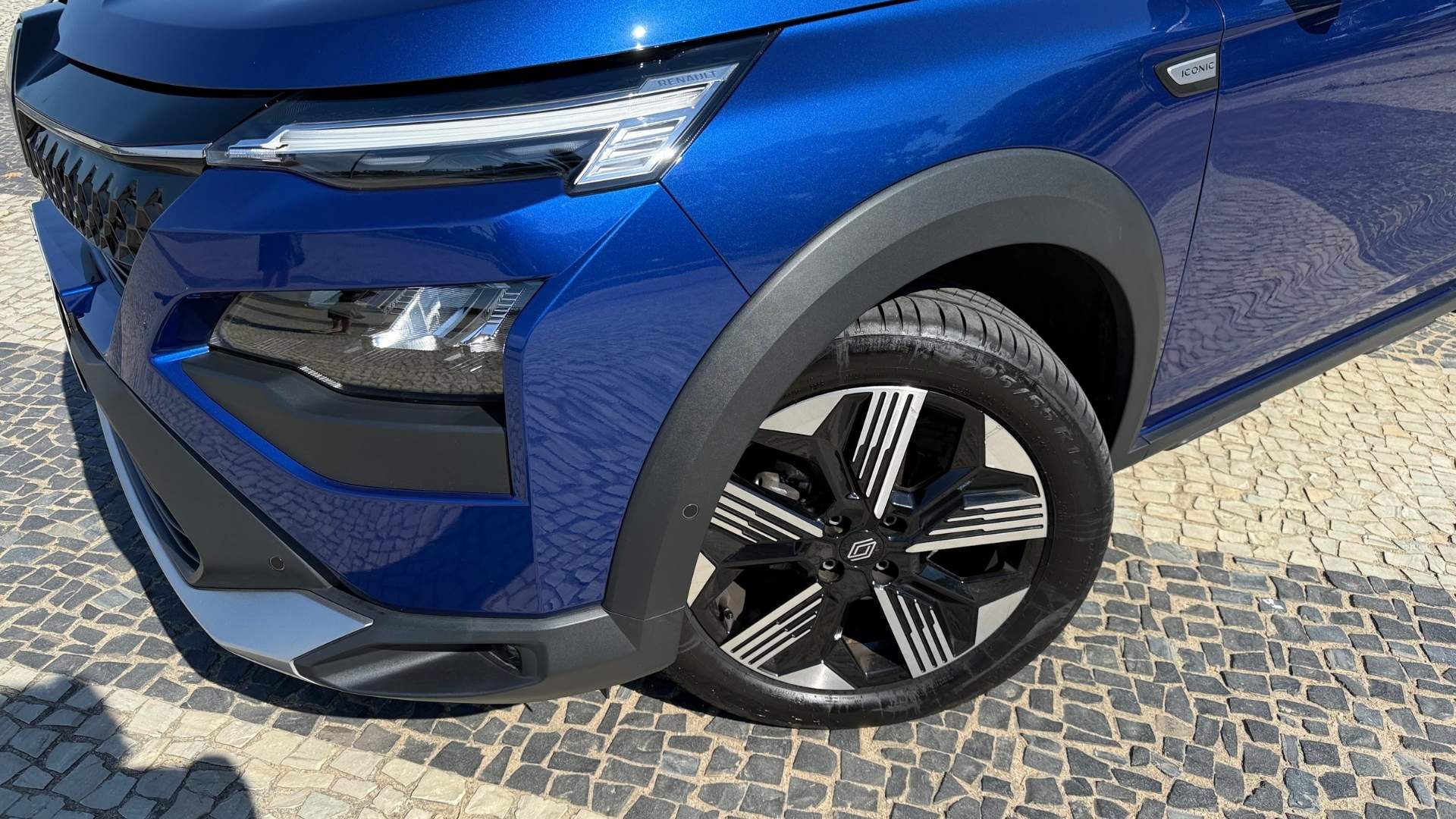 Close-up of the front left tire and wheel of a blue vehicle, showcasing the alloy wheel design and tire tread on a cobblestone pavement.