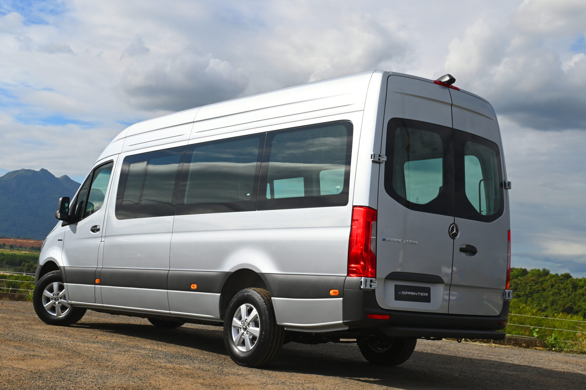 A silver Mercedes-Benz Sprinter van parked on a gravel surface with mountains in the background and partly cloudy skies.