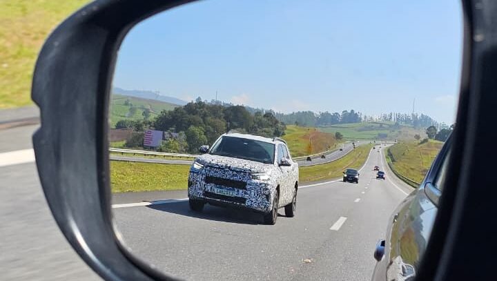 A view from a side mirror showing a camouflaged SUV on a highway, with green hills in the background.