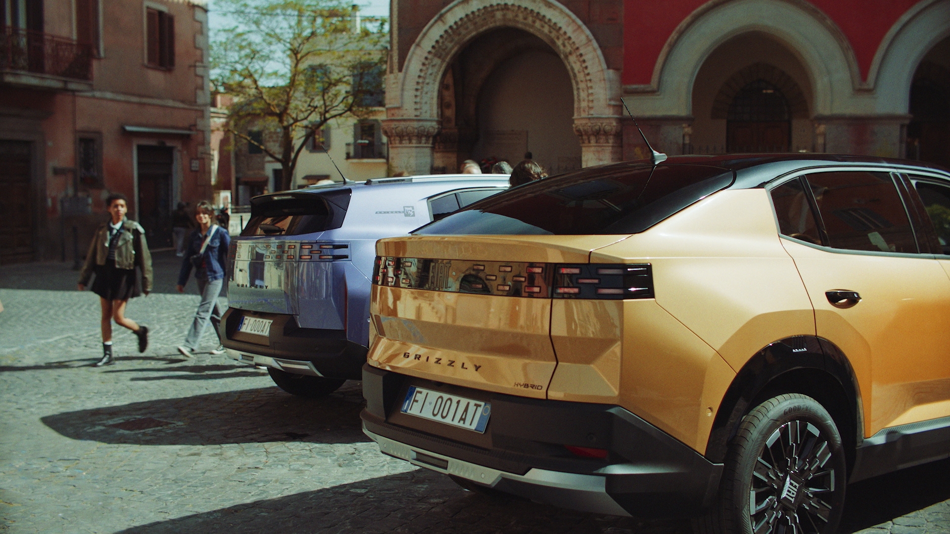 Two modern cars parked on a cobblestone street, with pedestrians walking in the background. The car on the left is a light blue color and the one on the right is a metallic gold, both featuring unique rear designs.