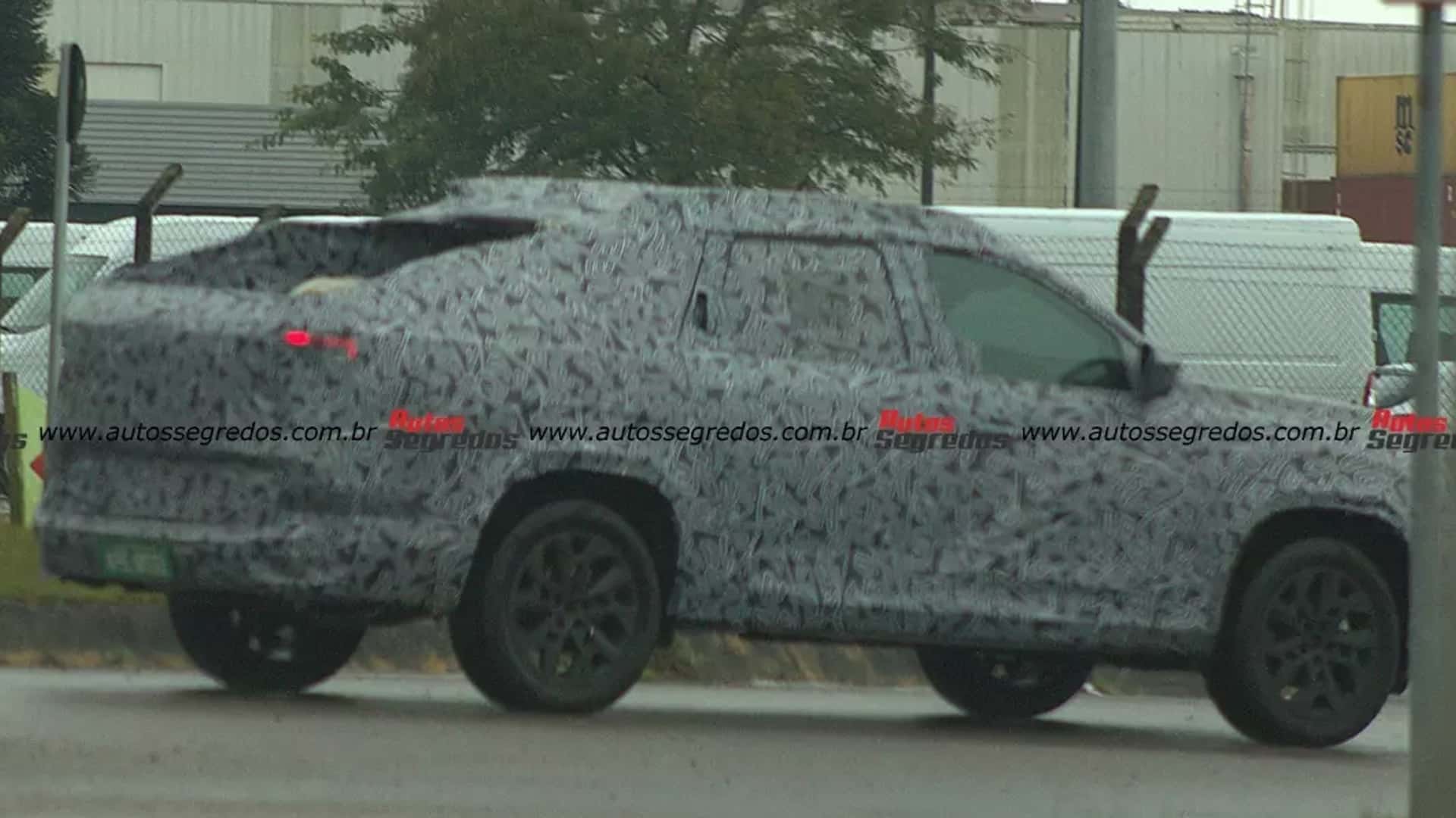 A camouflaged pickup truck is seen parked on a street, with blurred background featuring trees and industrial buildings.