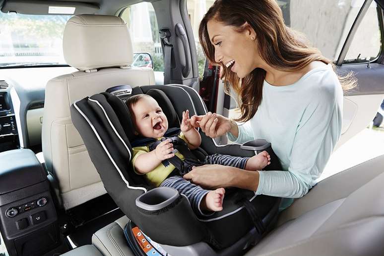 Uma mãe sorrindo e brincando com seu bebê em uma cadeirinha de carro, dentro de um veículo.