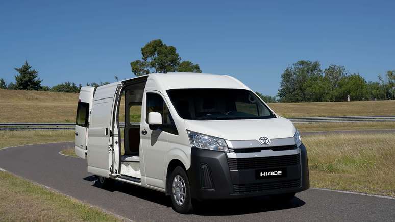 Furgão Toyota HiAce branco estacionado em uma estrada com grama ao fundo e céu azul.