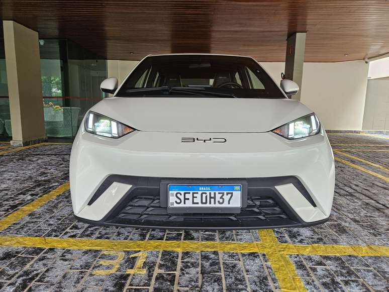 Carro elétrico branco da marca BYD estacionado em um estacionamento, com foco na parte frontal e placa de identificação visível.