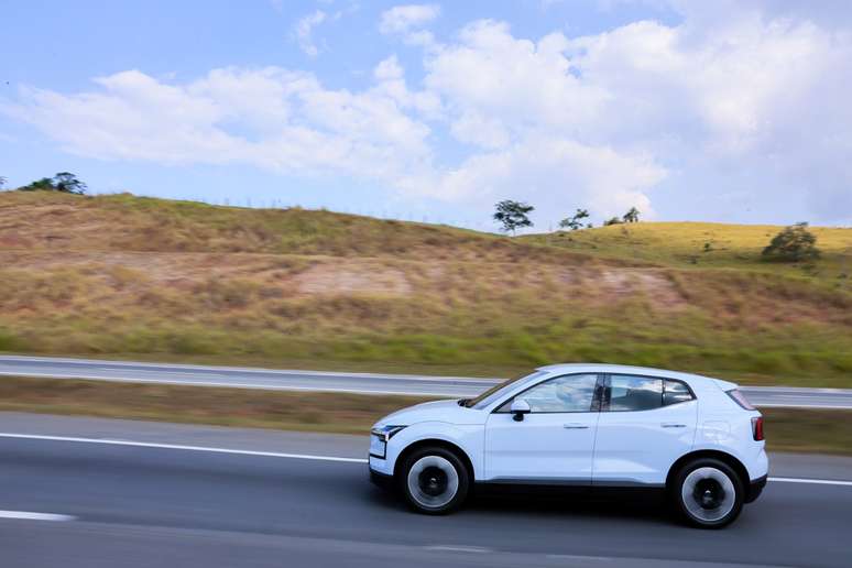Um carro branco se desloca rapidamente em uma estrada, com um cenário de colinas e céu azul ao fundo.