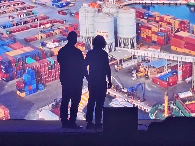Silhuetas de duas pessoas observando um porto com contêineres coloridos e estruturas industriais ao fundo.