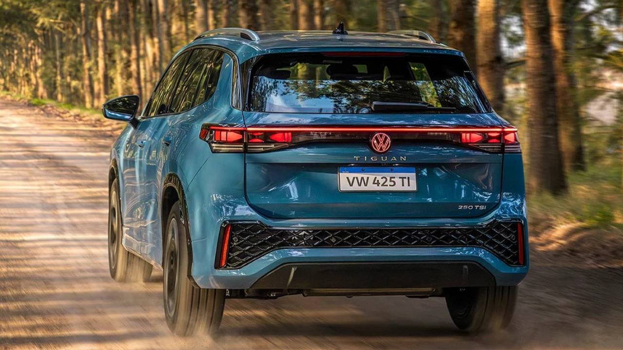 Um carro SUV Volkswagen Tiguan azul visto de trás, rodando em uma estrada de terra cercada por árvores.