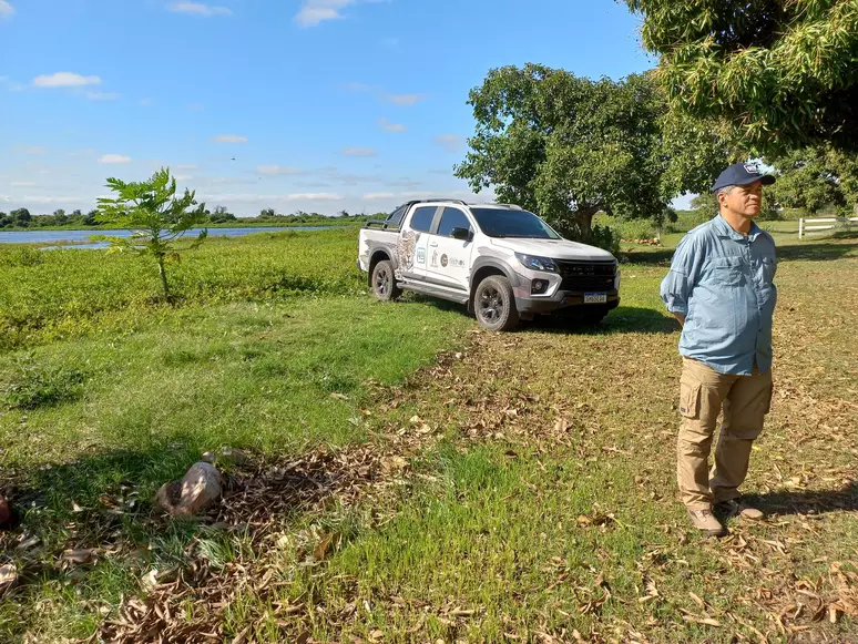 Homem em pé na natureza, ao lado de uma caminhonete branca estacionada, com um lago ao fundo e vegetação ao redor.
