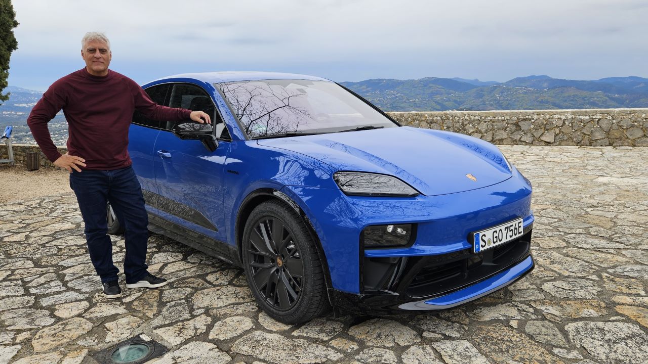 Um homem em pé ao lado de um carro elétrico azul em um local ao ar livre, com uma paisagem montanhosa ao fundo.