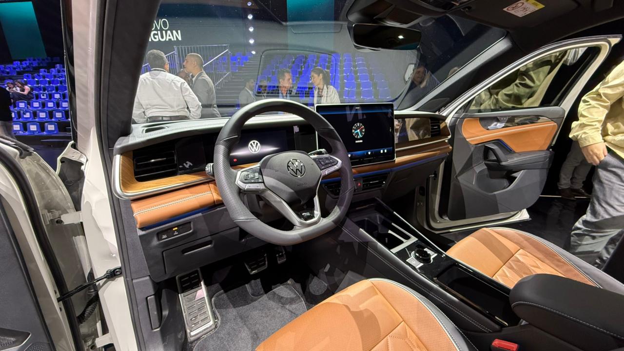 Interior de um carro Volkswagen, destacando o volante, painel com tela digital e acabamentos em couro marrom na cabine.