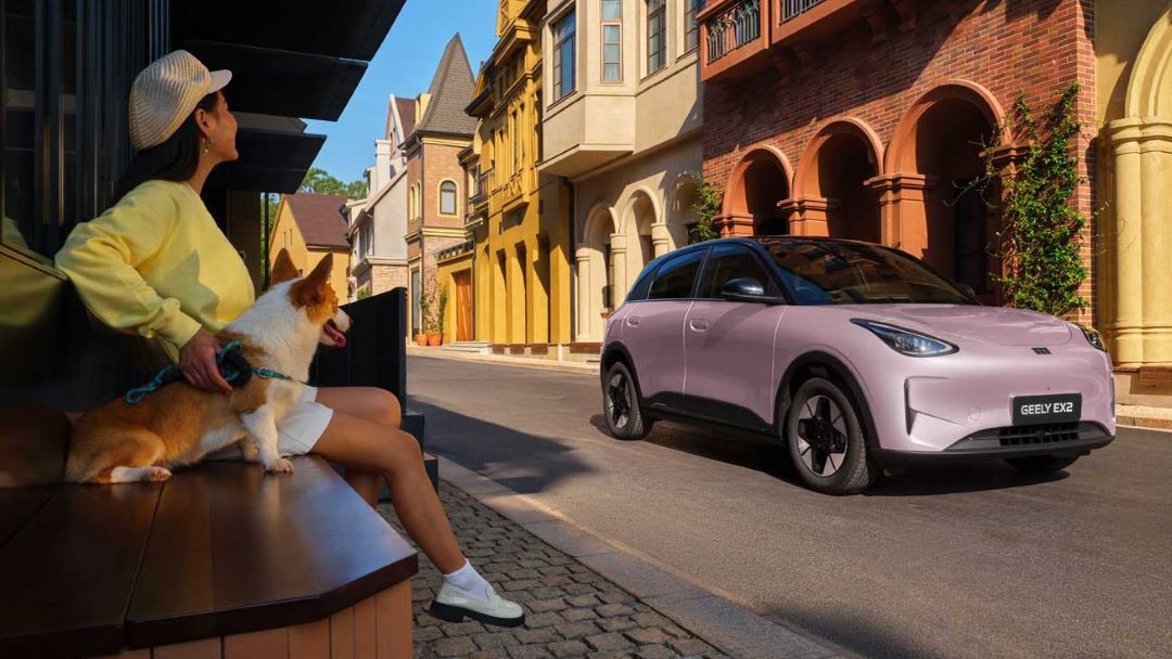 Uma mulher sentada em um banco de madeira, segurando a coleira de um cachorro da raça corgi, em uma rua com casas coloridas e um carro elétrico rosa estacionado.
