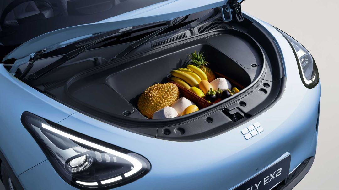 Imagem do porta-malas de um carro elétrico azul, com uma cesta contendo frutas como abacaxi, bananas, durião e frutas diversas.