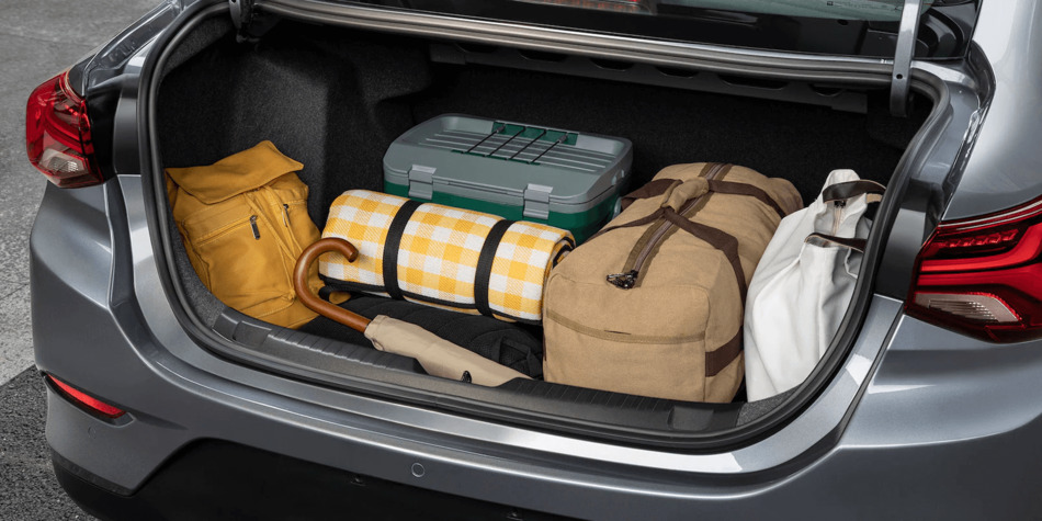 Malas e itens organizados no porta-malas de um carro, incluindo uma mochila amarela, uma cesta de piquenique com padrão quadriculado, uma bolsa de viagem grande e uma caixa cinza.