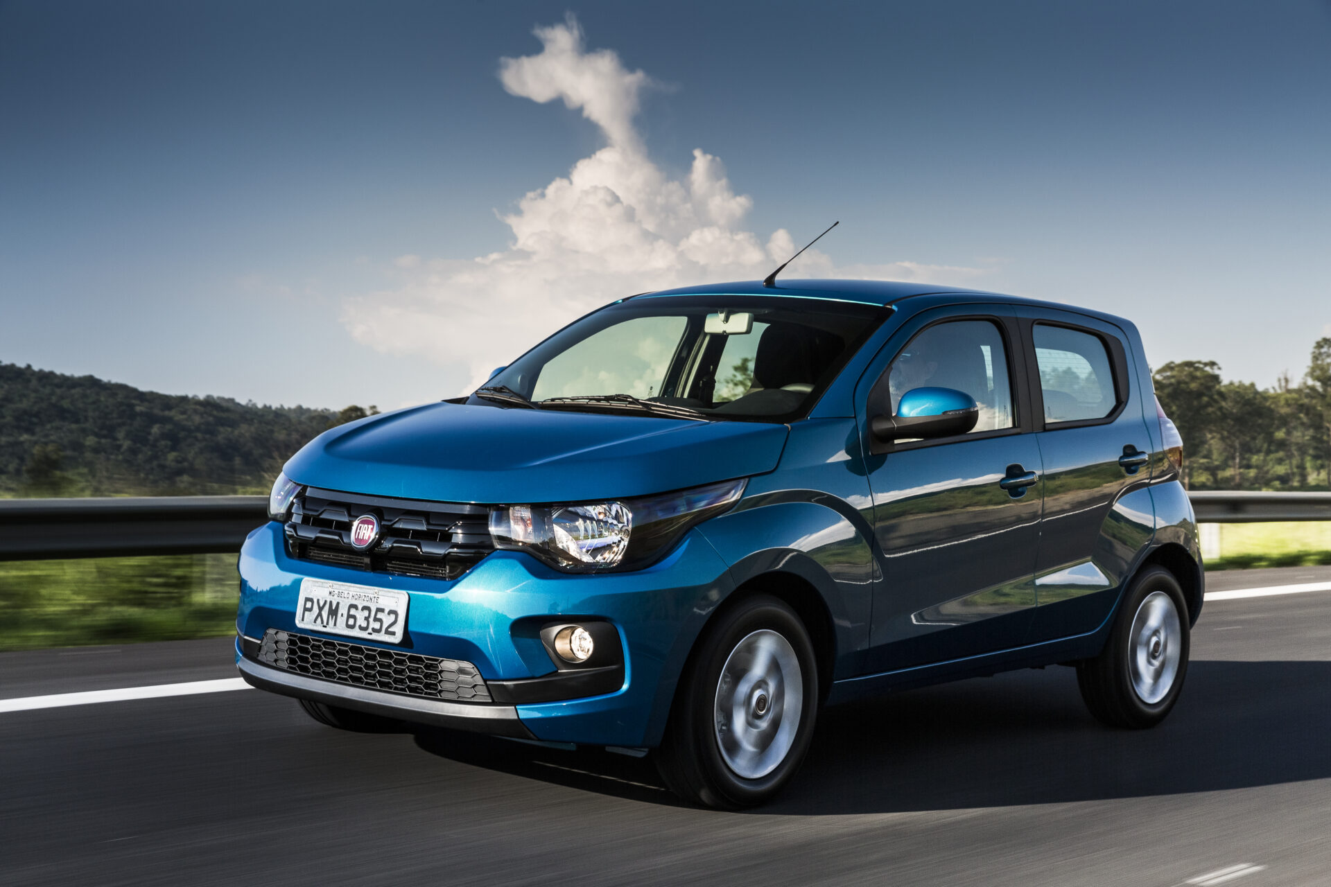 Carro da marca Fiat na cor azul, em movimento em uma estrada cercada por natureza e céu limpo.