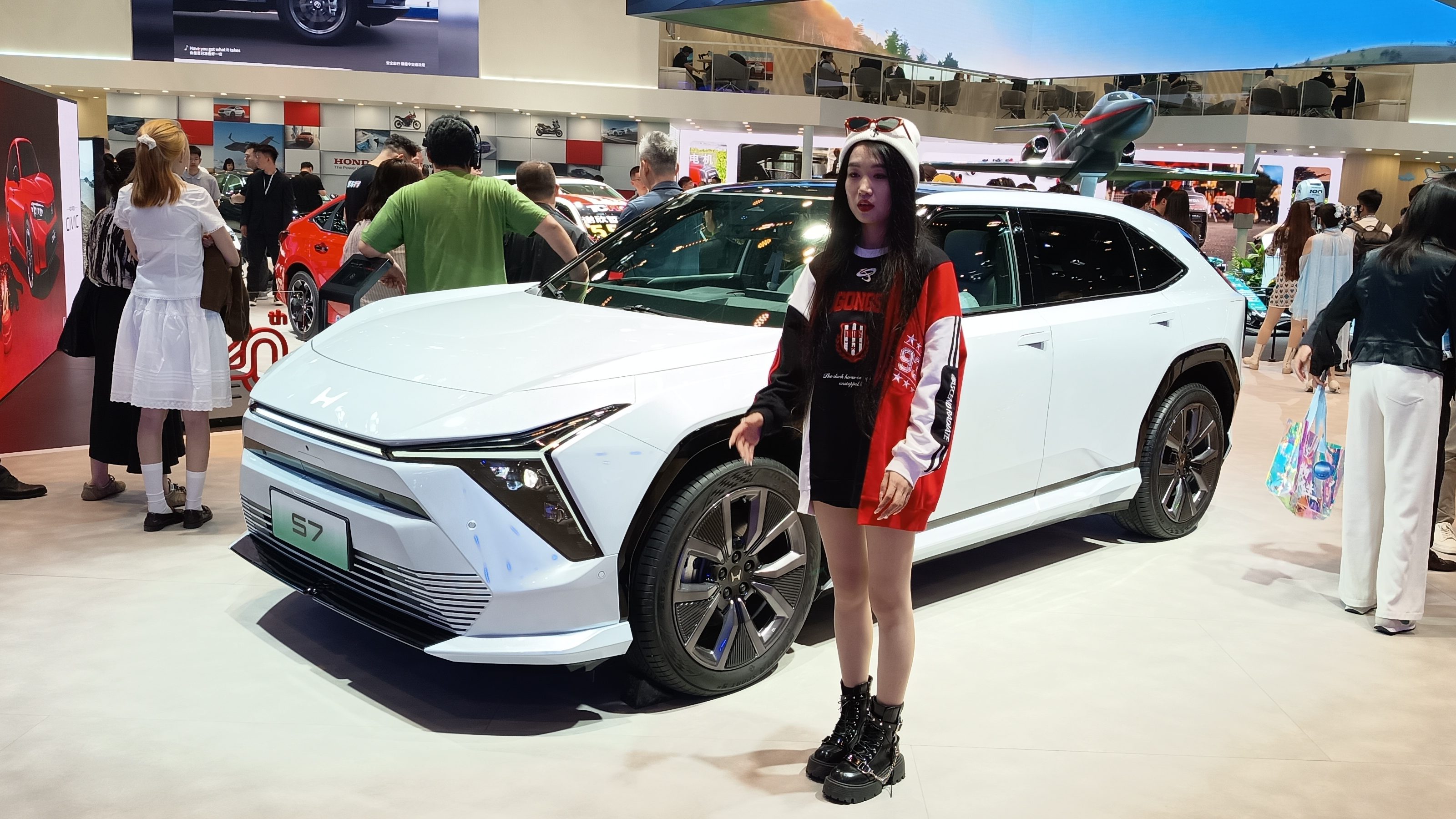 Uma jovem em pé ao lado de um carro elétrico branco em um salão do automóvel, com pessoas ao fundo observando outros veículos.