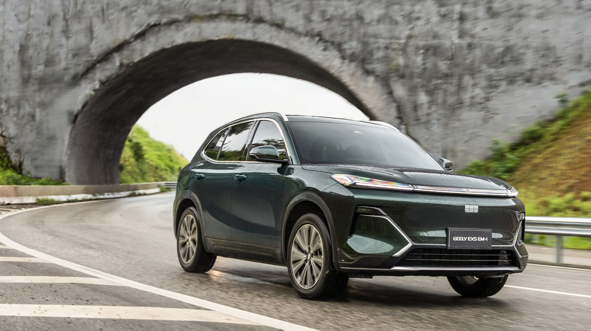 SUV em movimento em uma estrada curvilínea sob um túnel, com paisagens verdes ao fundo.