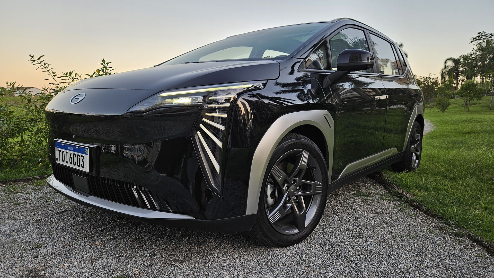 Um carro elétrico preto estacionado em um caminho de cascalho, com um fundo de grama e árvores ao entardecer.