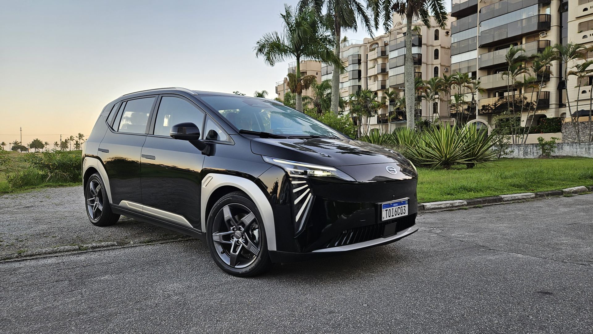 Um carro elétrico preto em uma rua com prédios ao fundo e palmeiras nas laterais.