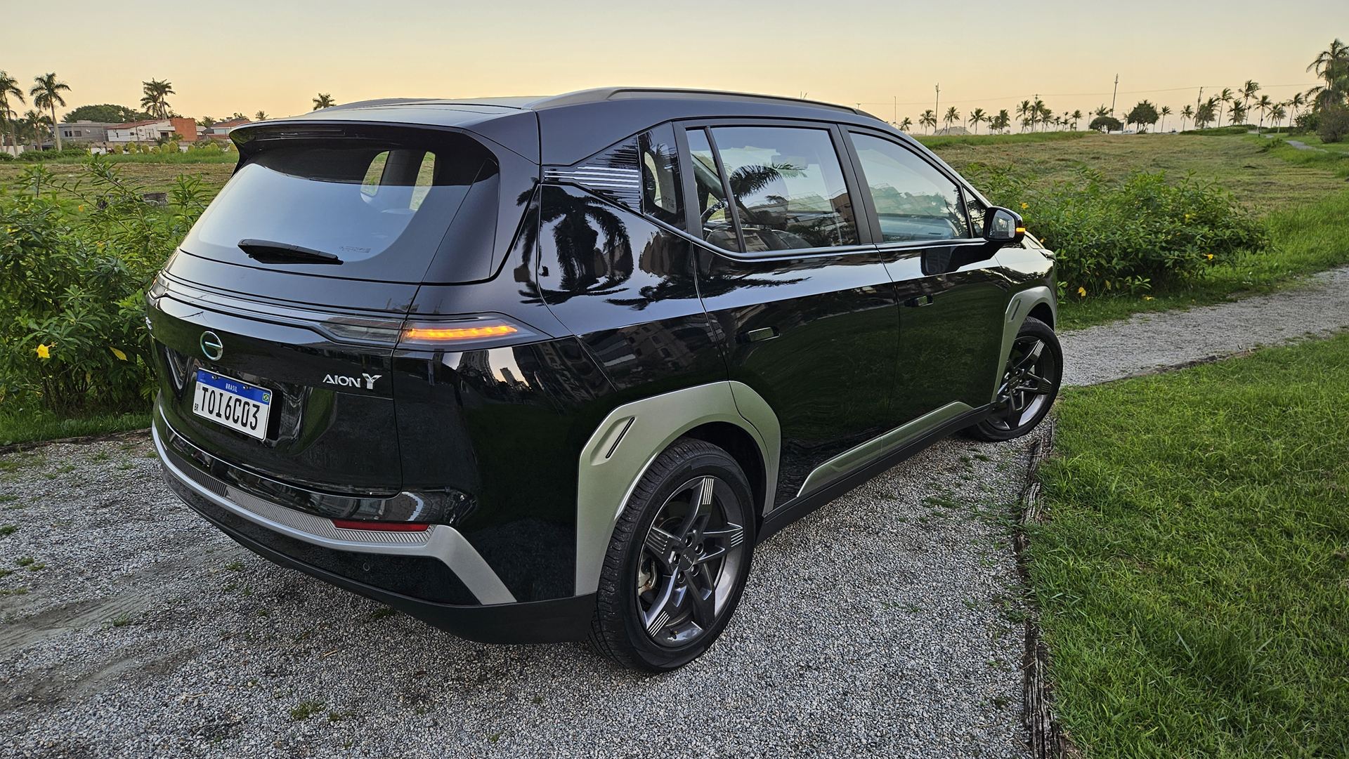 Um carro elétrico modelo Aion Y preto estacionado em um caminho de cascalho, com um fundo de grama verde e palmeiras ao fundo.