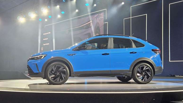SUV azul em destaque em um palco iluminado, com fundo escuro e painéis gráficos ao fundo.