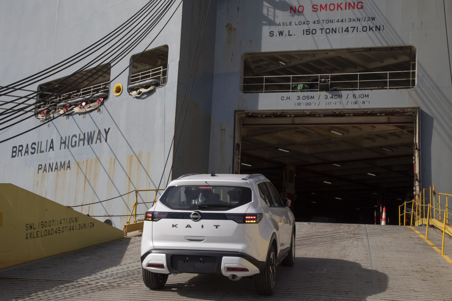 Um carro branco está saindo de um navio cargueiro chamado Brasília Highway, que está atracado no porto.