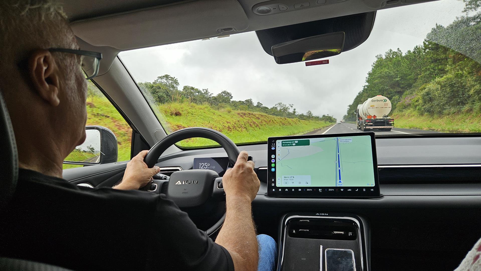 Pessoa dirigindo um carro, com as mãos no volante e olhando para a estrada à frente. Um painel digital com mapas é visível ao lado.
