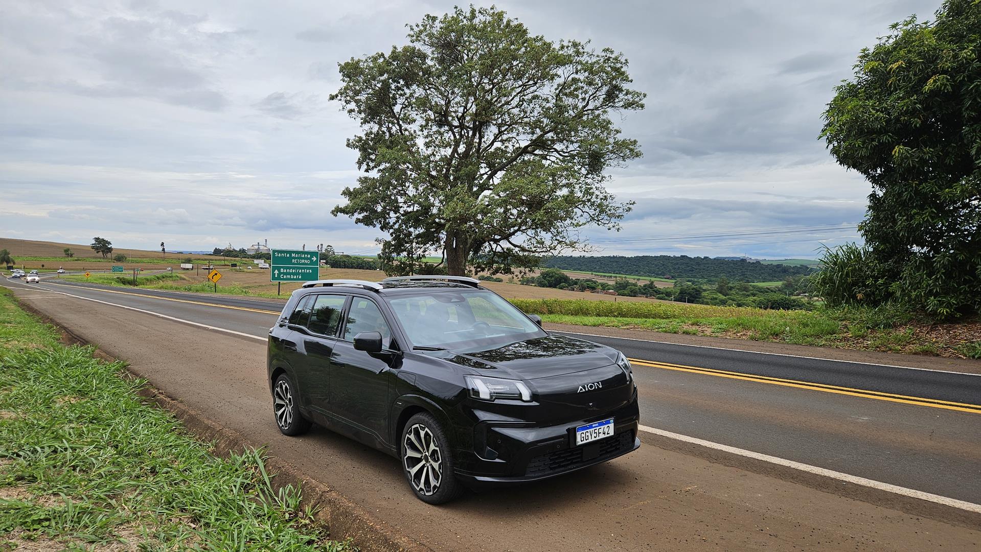 Um carro preto estacionado ao lado de uma estrada, com placas de sinalização indicando direções. Ao fundo, há uma paisagem com árvores e um céu nublado.