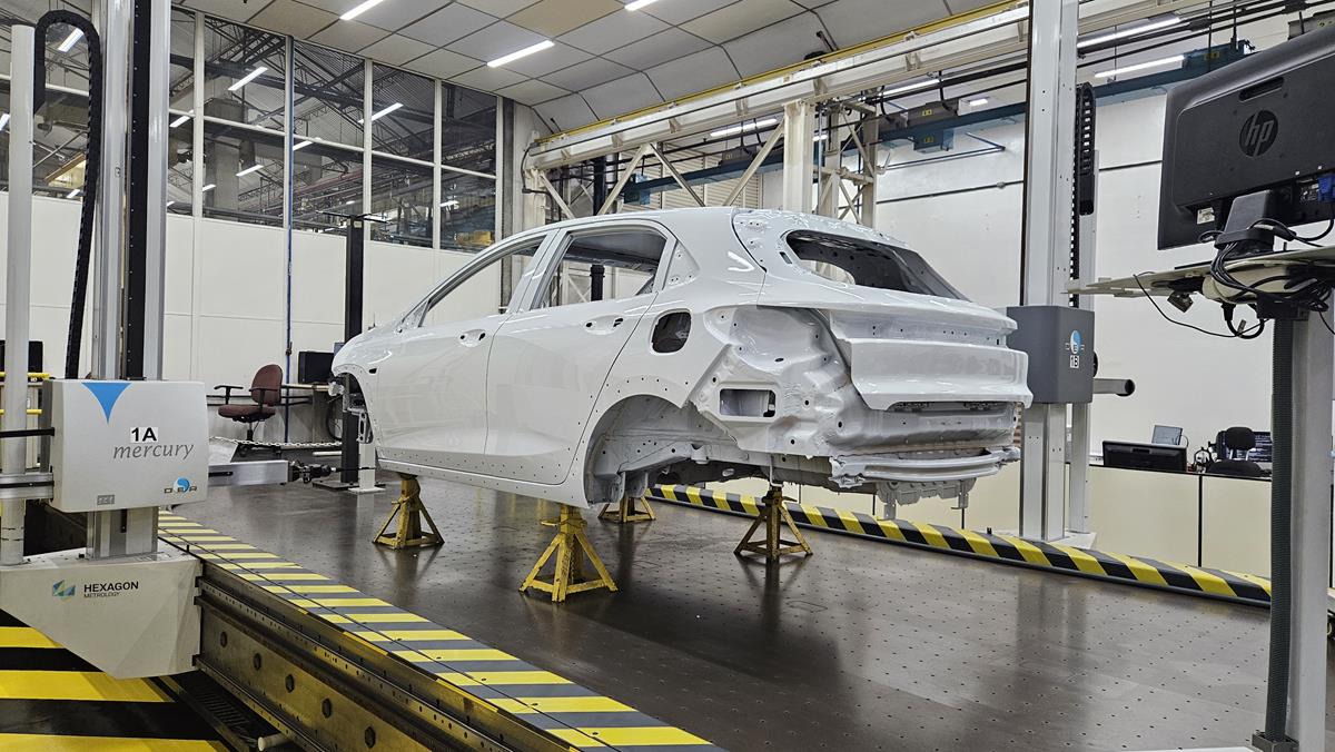 Estrutura de um carro branco em uma linha de produção, com equipamentos de medição ao fundo.