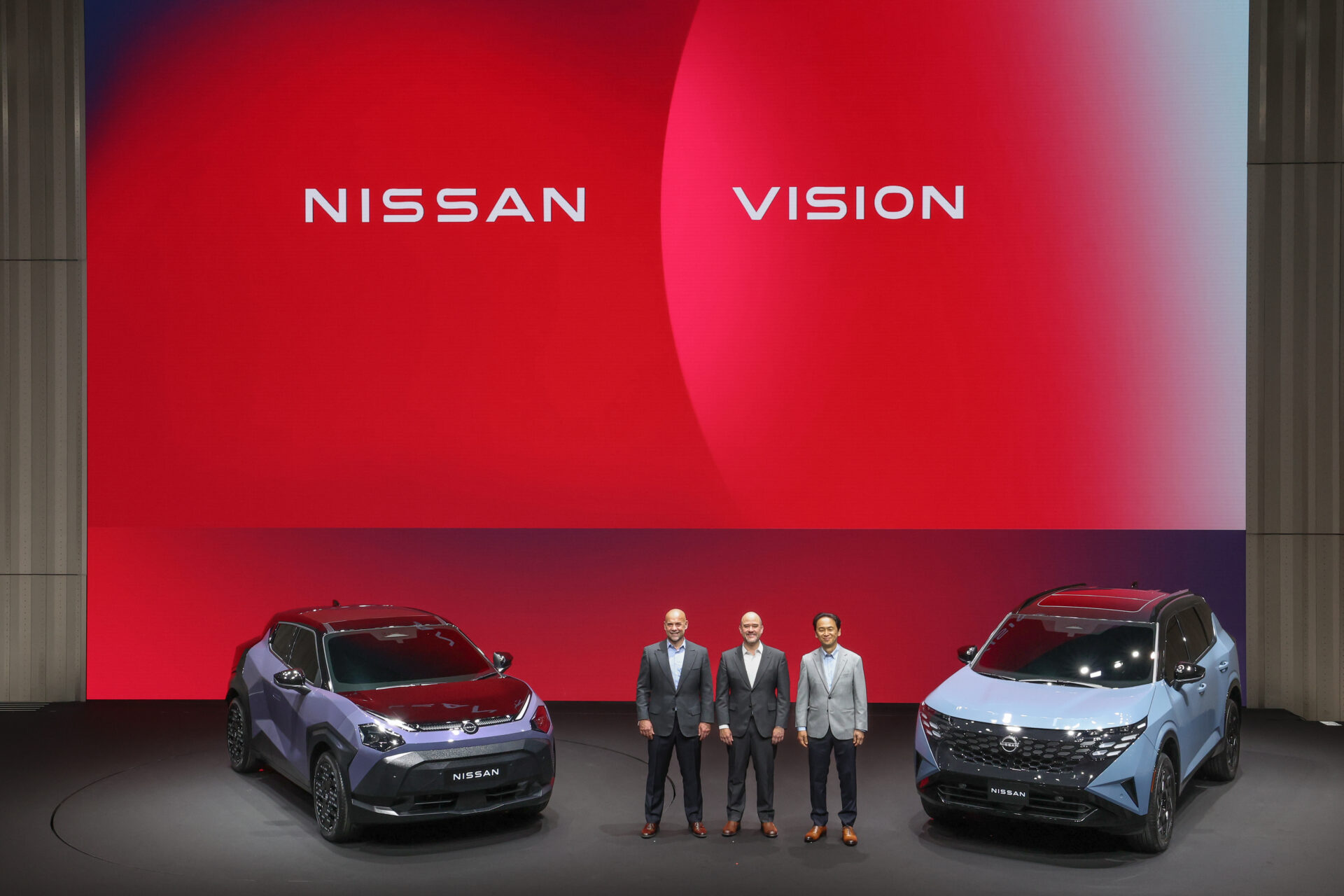 Apresentação de novos modelos da Nissan em um evento, com dois carros ao lado de três homens em trajes formais, em frente a um fundo colorido com as palavras 'Nissan Vision'.
