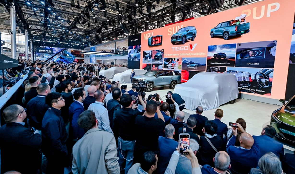 A large crowd of people at an automotive exhibition, capturing photos of covered vehicles on stage, with a prominent display showcasing the Tiggo V model and features.