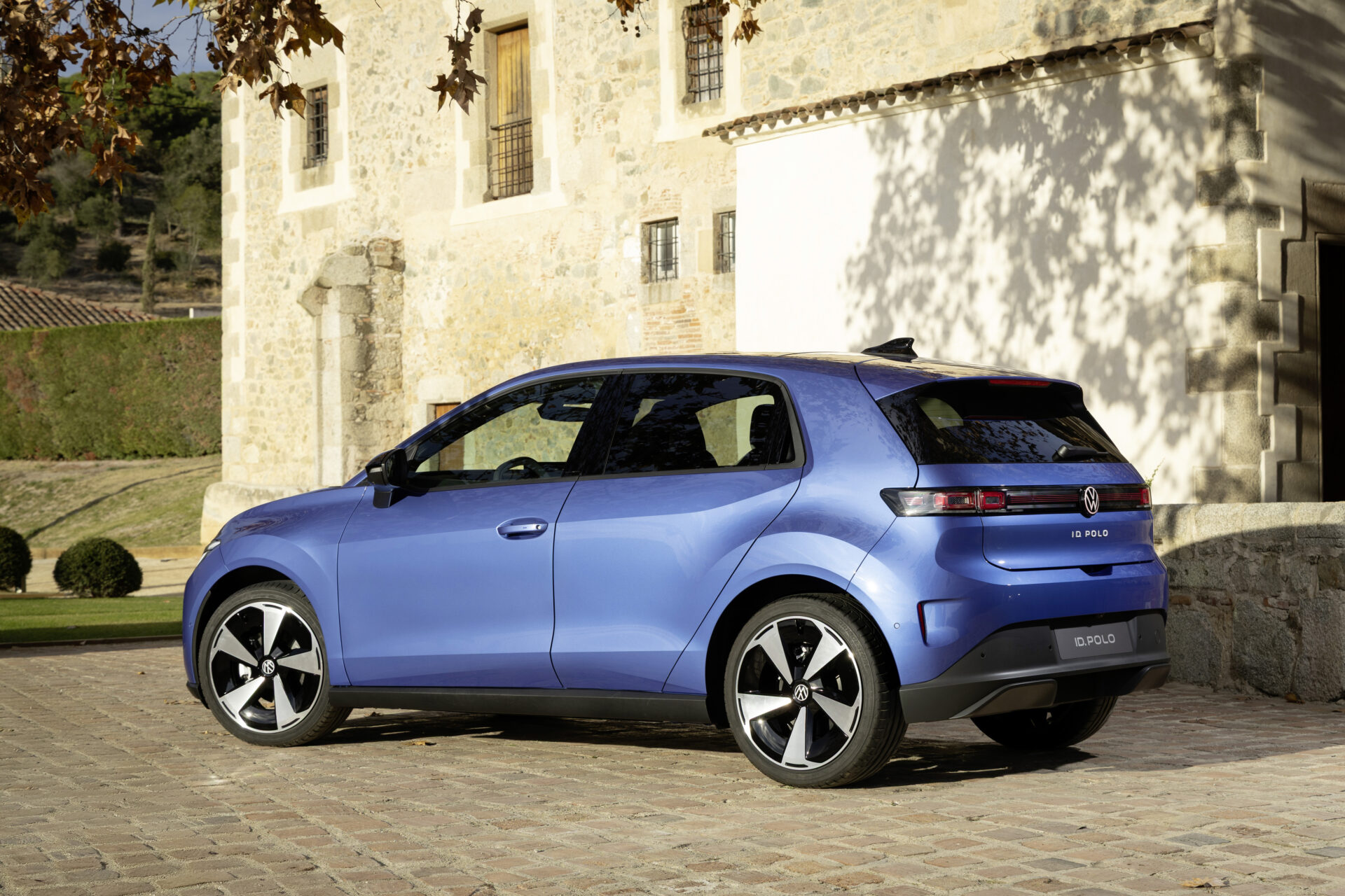 Um carro compacto Volkswagen ID. Polo azul estacionado em uma calçada de pedras, com uma construção de pedra ao fundo.