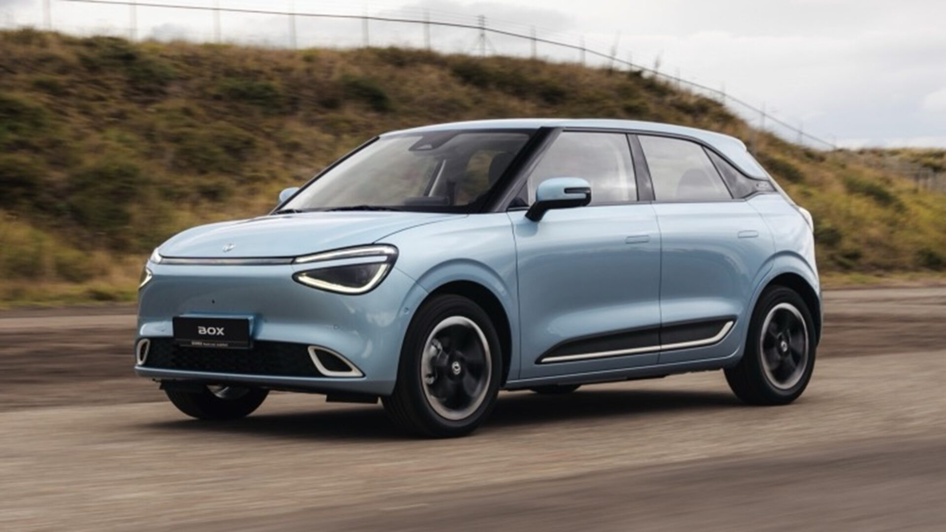 A blue compact SUV in motion on a road with a grassy hillside in the background.