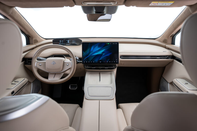 Interior de um carro moderno com volante e painel de controle elegante, incluindo uma tela grande no centro e acabamentos em tons de bege.