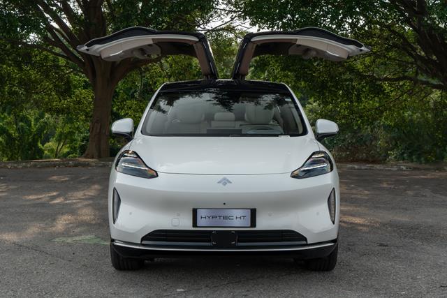 Vista frontal de um carro elétrico branco com portas tipo asa de gaivota abertas, estacionado em um ambiente com árvores ao fundo.