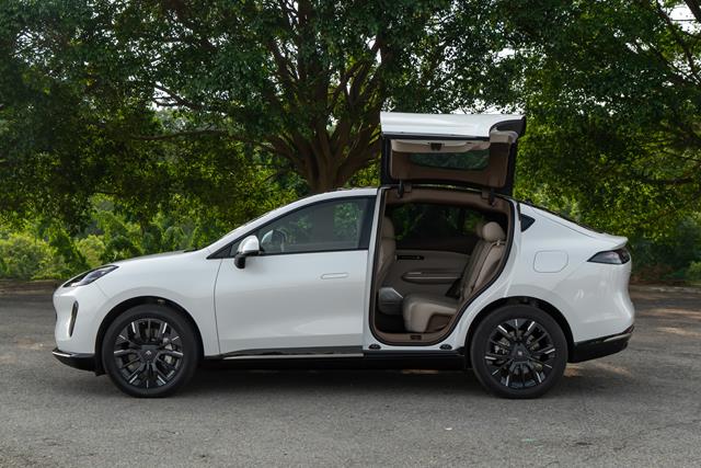 Um carro branco visto de lado, com a porta traseira levantada, estacionado em uma área arborizada.
