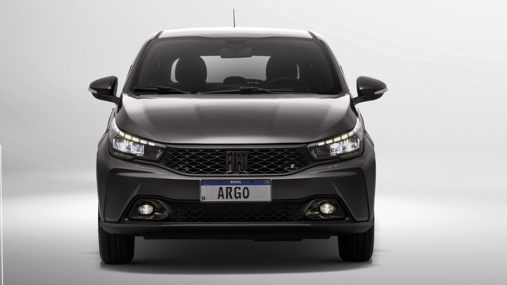 Frente de um carro Fiat Argo em fundo claro, destacando a grade frontal e os faróis iluminados.