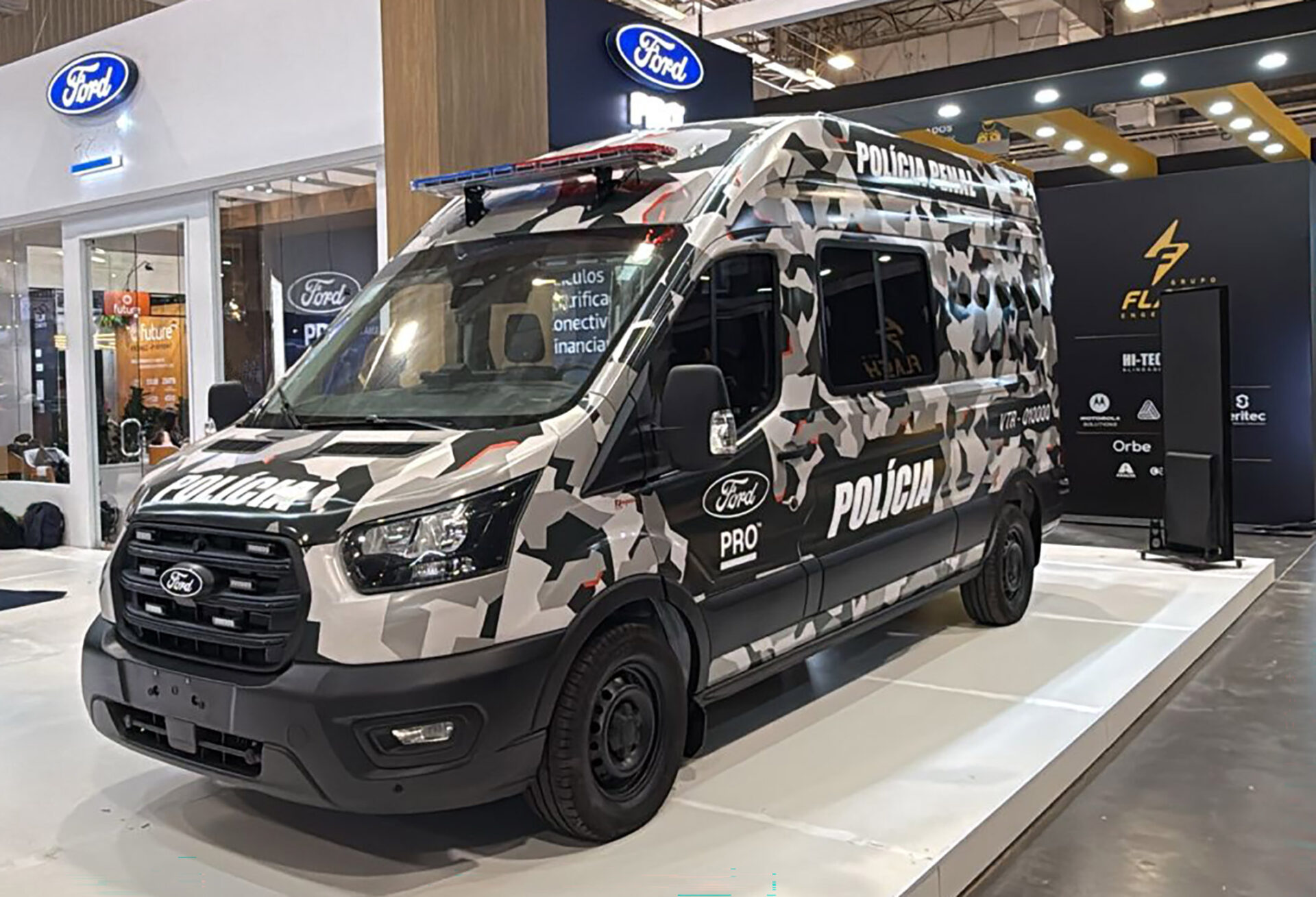 Furgão policial da Ford exibido em um evento, com pintura camuflada e luzes de emergência no topo.