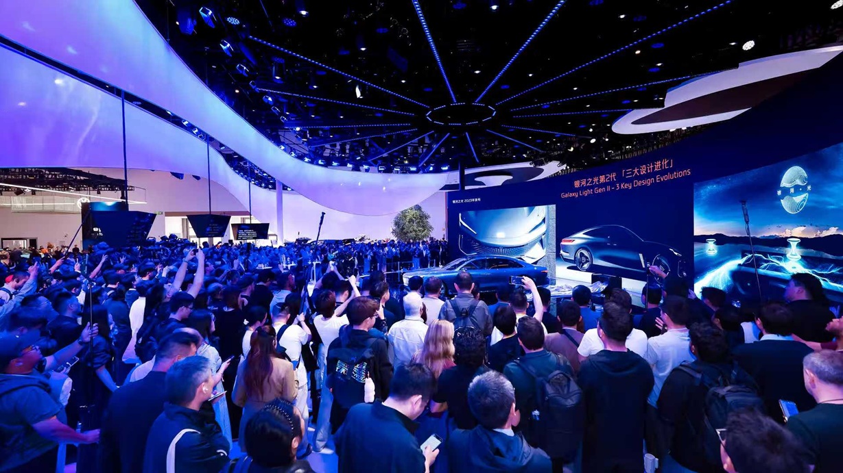 Crowd gathered around a futuristic car display at an automotive exhibition, with vibrant blue lighting and digital screens showcasing design elements.
