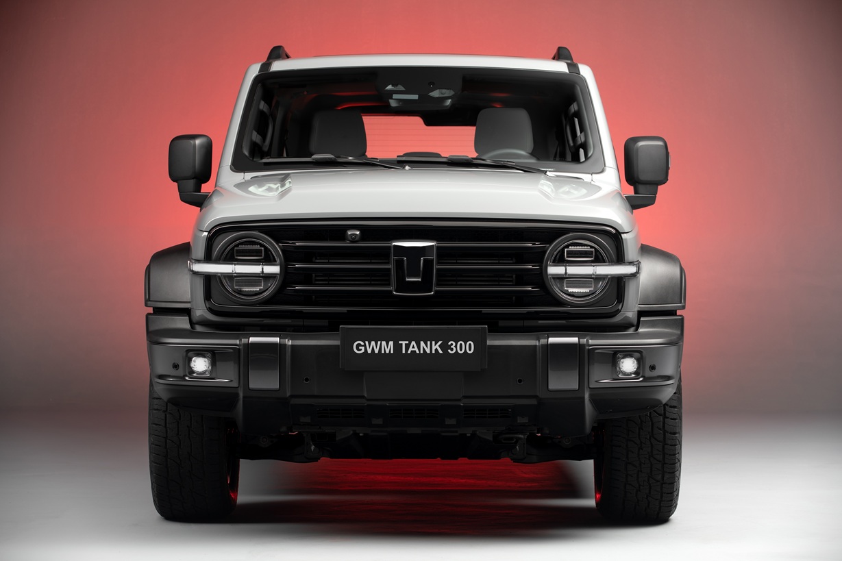 Front view of a GWM Tank 300 SUV with a sleek design, featuring a bold grille and distinctive headlights against a red and gray background.