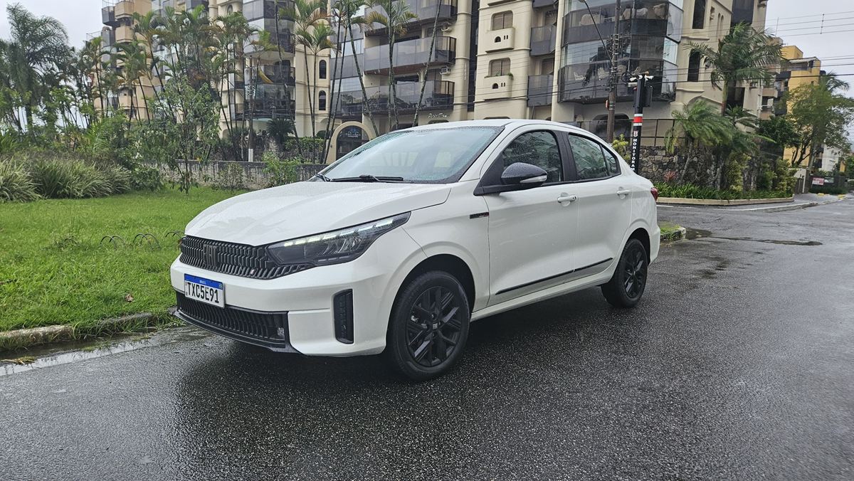 Um carro sedã branco estacionado em uma calçada, com árvores e prédios ao fundo em um dia chuvoso.