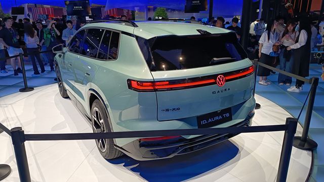 Vista traseira do carro elétrico Volkswagen ID.Aura T6 em uma exposição, com público ao fundo.