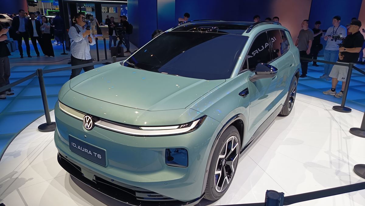 Carro elétrico Volkswagen ID. Aura T6 em exposição, com design moderno e cor verde, cercado por visitantes em um evento.