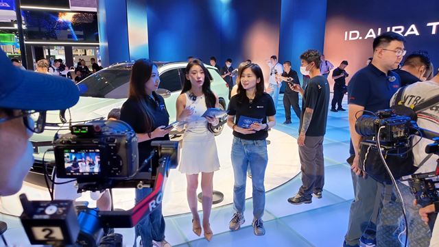 Duas apresentadoras conversando em um evento com um carro ao fundo, enquanto são filmadas por várias câmeras, com uma audiência ao redor.