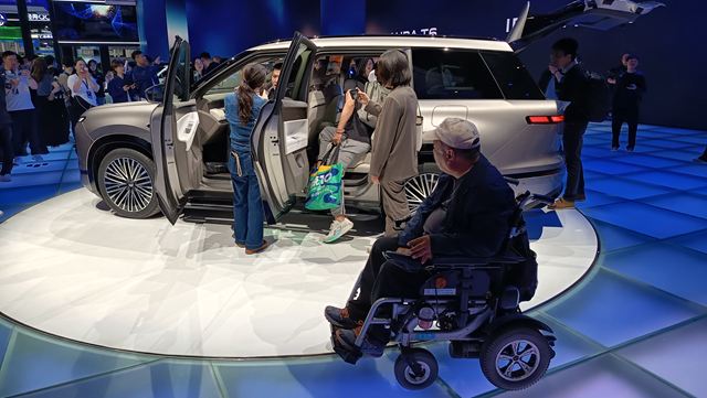Grupo de pessoas interagindo com um carro SUV em um evento, com um homem em cadeira de rodas observando.