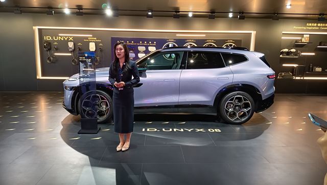 Uma mulher em traje profissional apresenta o carro elétrico ID. UNYX 08 em uma exposição, com um cenário moderno ao fundo.