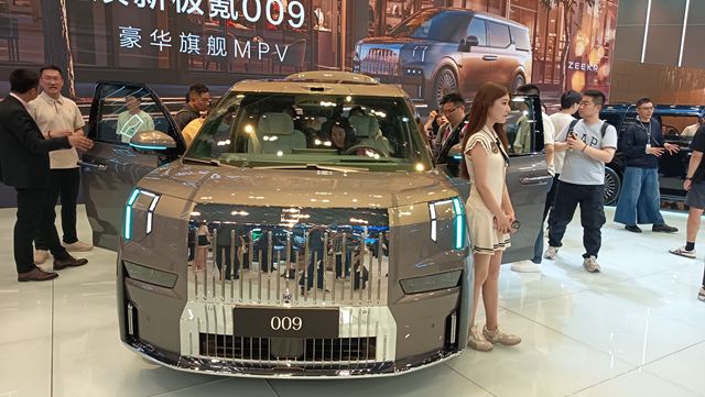 Uma exposição de automóveis mostra um veículo modelo 009 com design moderno e detalhes cromados. Pessoas estão reunidas ao redor do carro, enquanto uma jovem mulher posa ao lado dele.